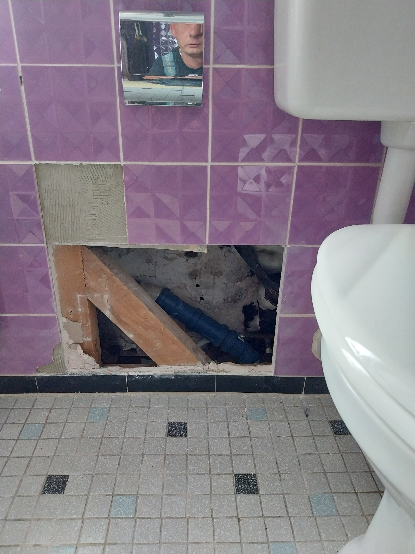 Wand mit fehlenden Fliesen, sichtbarem Rohrschacht und Reparaturbereich hinter der Toilette